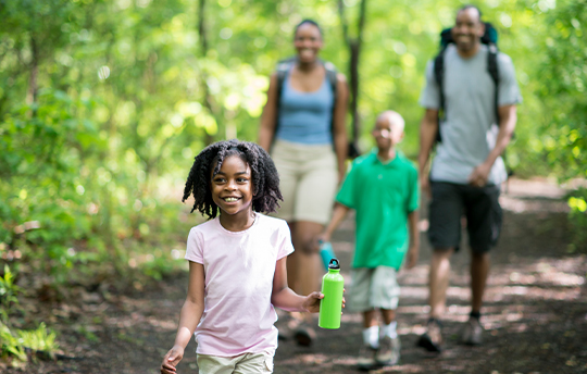 African-American family hiking in woods, with little girl walking in font smiling with water bottle.